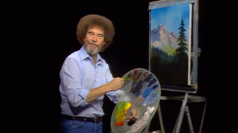 Bob Ross sitting on chair with painting by his side.